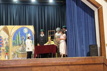 A színpadon 6 szereplő, a király ül az asztalnál koronával a fején, a többiek körülötte állnak.