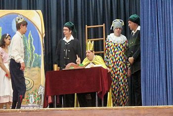 A színpadon a király ül az asztalnál mutatóújját felemelve, körülötte négyen állnak.